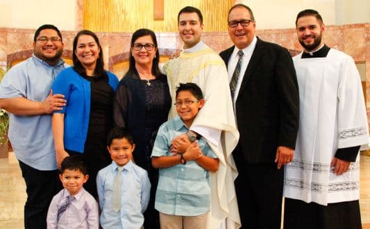 family with adults and kids standing next to priest.